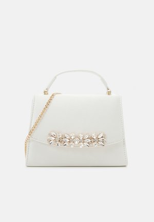 Kleine weiße Handtasche mit kurzem Henkel, goldener Kettenschulterriemen und juwelenbesetzter Blumenverzierung auf der Frontklappe, vor einfachem Hintergrund.