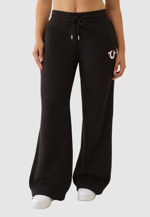 Pantalons de survêtement noirs avec une taille à cordon, jambes larges, et un accent logo rose. Fabriqués en tissu doux avec une texture lisse.