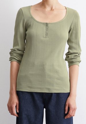 Schmal geschnittenes, geripptes, olivgrünes Langarmshirt mit Rundhalsausschnitt und drei Knöpfen auf der Brust, getragen zu dunkelblauen Jeans.