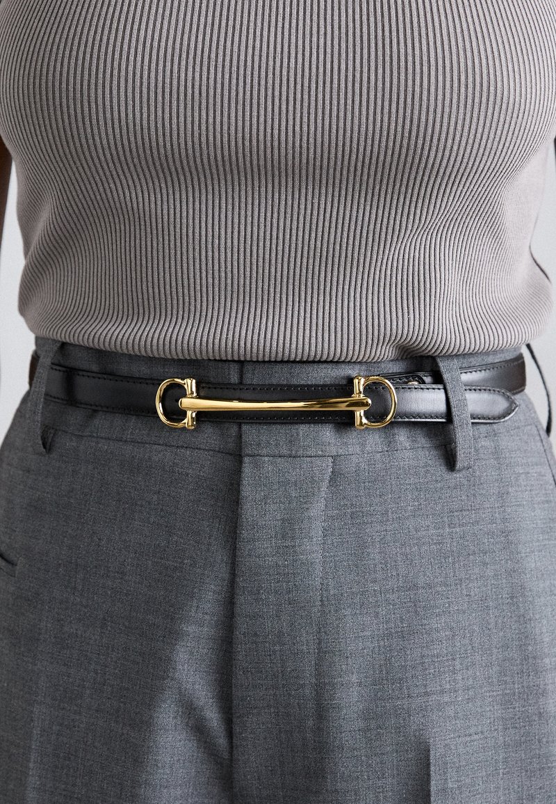 Black leather belt with a gold circular buckle and bar detail, worn over gray patterned trousers. Ribbed gray top visible above.