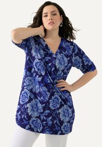 Kvinna iklädd en blå blommig wrap-top med korta ärmar och vita byxor, poserande med en hand på halsen och den andra på höften.