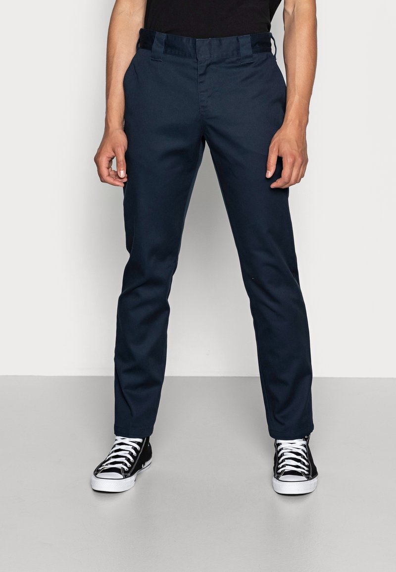 Personne portant un pantalon droit bleu foncé et des baskets montantes noires avec des lacets blancs, debout devant un fond uni.