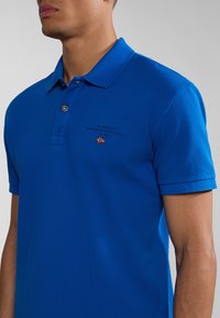 Polo shirt blu realizzato in tessuto testurizzato, con colletto a due bottoni, maniche corte arrotolate e un piccolo logo con un accento a forma di bandiera.