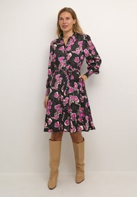 Schwarzes Kleid mit pinkem Blumenmuster, lange Ärmel, Taillengürtel, ausgestellt Rock, kombiniert mit beigen kniehohen Stiefeln. Glattes Material.