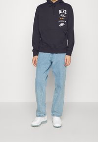 Svart huvtröja med "Nike"-logotyp i vitt och orange, känguruficka, ribbade muddar, parat med ljusblå jeans och vita sneakers.