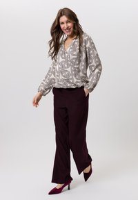 Blouse met patroon in abstracte vormen in beige en bruin, gecombineerd met dieprode bordeauxrode broek en bijpassende pumps met een spits uiteinde, getoond in beweging.