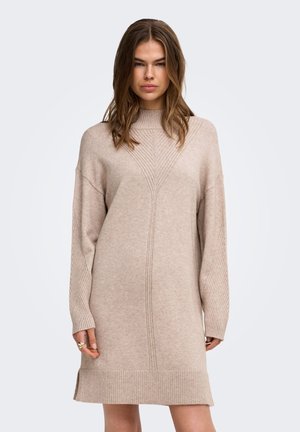 Femme aux longs cheveux bruns porte une robe pull beige en tricot avec des poignets et un ourlet côtelés, debout devant un fond clair uni.