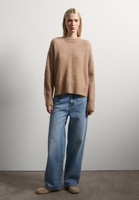Blonde Frau, die einen lockeren beigen Pullover, hellblaue weite Jeans und beige Schuhe trägt und vor einer einfachen weißen Wand steht.