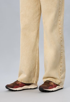 Persona con jeans larghi color crema e sneakers marroni con suola color crema, in piedi su pavimento grigio.