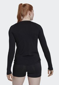 Frau mit roten Haaren im Pferdeschwanz, die ein langärmliges schwarzes Oberteil und schwarze Shorts trägt, steht mit dem Rücken zu einem einfarbigen Hintergrund.
