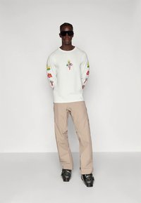 Fehér hosszú ujjú ing grafikai mintákkal az ujjain, bézs nadrág és fekete napszemüveg, sima háttér előtt állva.