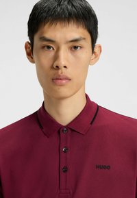 Burgunderfarbendes Poloshirt mit Kragen, drei Knöpfen und schwarzen Akzenten. Mit kleinem schwarzem Logo auf der Brust. Strukturierter Stoff.