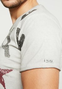 Camiseta de algodón gris con gráficos impresos en negro y rojo, mangas cortas y una pequeña etiqueta "Key Largo" en la manga. Textura suave.