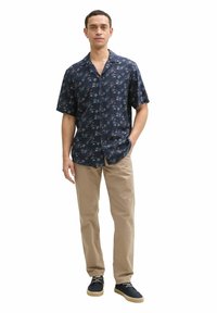 Kurzärmliges, navy-farbenes Hemd mit Knopfleiste und Palmenmuster. Kombiniert mit hellbeigen Hosen und navy-farbenen Schnürschuhen.