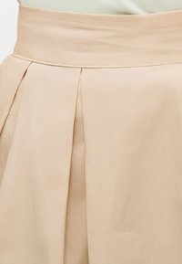 Μπεζ φούστα με ψηλή μέση και πιέτες μπροστά, εμφανίζεται από τη μέση μέχρι περίπου το μέσο μηρού, συνδυασμένη με λευκή μπλούζα με ριμπ ύφανση.