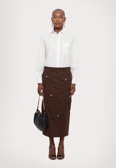 Weißes Langarmhemd mit einer Brusttasche, kombiniert mit einem braunen Midirock, der mehrere aufgesetzte Taschen und goldene Knöpfe hat. Schwarze Tasche und Absatzschuhe.