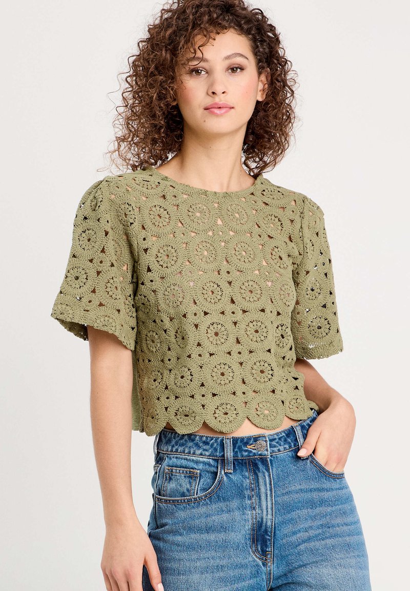 Top en crochet vert olive présentant des motifs circulaires, des manches courtes bouffantes et un ourlet festonné, associé à un jean en denim taille haute.