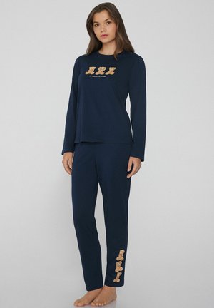 Vrouw in een marineblauwe lange mouwen pyjama set met teddybeerontwerpen en de tekst "My Weekly Attitude", staand op blote voeten tegen een lichte achtergrond.