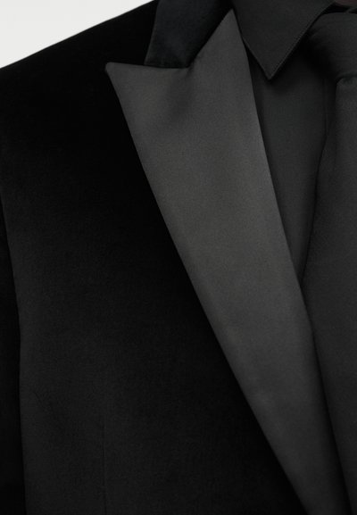 Μαύρο σακάκι βελούδου γιασημί με λείο σατέν γιακά αιχμηρής γραμμής, φορεμένο πάνω από μαύρο πουκάμισο και μαύρη γραβάτα.