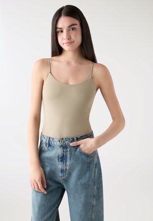 Giovane donna con capelli lunghi e scuri che indossa una canottiera beige e jeans blu a vita alta, in piedi con una mano in tasca davanti a uno sfondo semplice.