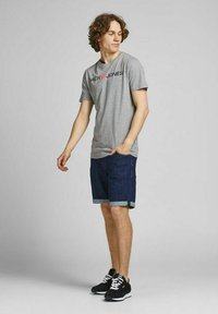 Graues Baumwoll-T-Shirt mit "Jack & Jones"-Logo, kombiniert mit blauen Jeansshorts mit umgeschlagenen Säumen und schwarzen Sportschuhen.