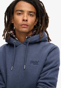 Felpa blu navy in un materiale morbido, con cappuccio regolabile, logo ricamato e un colletto a quadri visibile sotto.