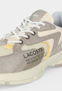 Μπεζ και κρεμ αθλητικό παπούτσι Lacoste με λευκά κορδόνια, υφασμένα πάνελ και εκτυπωμένη λεπτομέρεια συντεταγμένων στο πλάι.