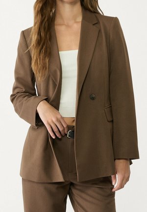 Femme portant un blazer marron et un pantalon assorti, un top court blanc côtelé, cheveux longs détachés, tenant le revers du blazer.