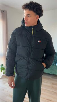 Musta toppatakki, jossa on huppu, keltainen vetoketju, sivutaskut ja pieni logo rinnassa. Yhdistetty vihreisiin housuihin.