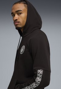 Mand iført en sort hættebluse med Manchester City-logo på brystet og mønstrede striber på ærmet, mod en grå baggrund.