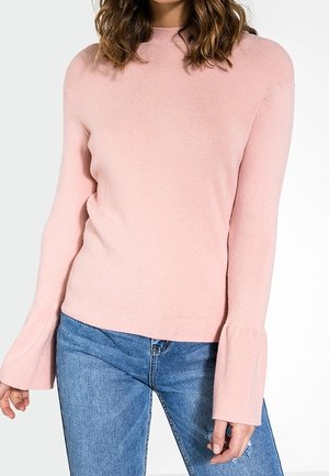 Maglione rosa a coste con colletto alto e maniche a campana, abbinato a jeans denim blu. Tessuto morbido e silhouette aderente.