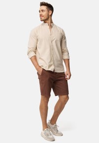 Chemise beige à manches longues avec un col montant, poche avant, associée à un short marron. Le modèle porte des baskets grises claires.