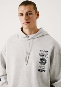 Sudadera gris hecha de un tejido suave con un bolsillo tipo canguro en la parte delantera. Presenta logotipos y texto negros de Red Bull Racing en el área izquierda del pecho.