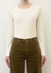 Langärmliges cremefarbenes Oberteil aus strukturiertem Stoff, kombiniert mit olivgrünen Cordhosen mit Knopfverschluss und Taschenverzierungen.