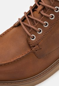 Scarpa in pelle marrone con punta rotonda, occhielli in metallo e lacci marroni; presenta cuciture visibili e una finitura testurizzata.