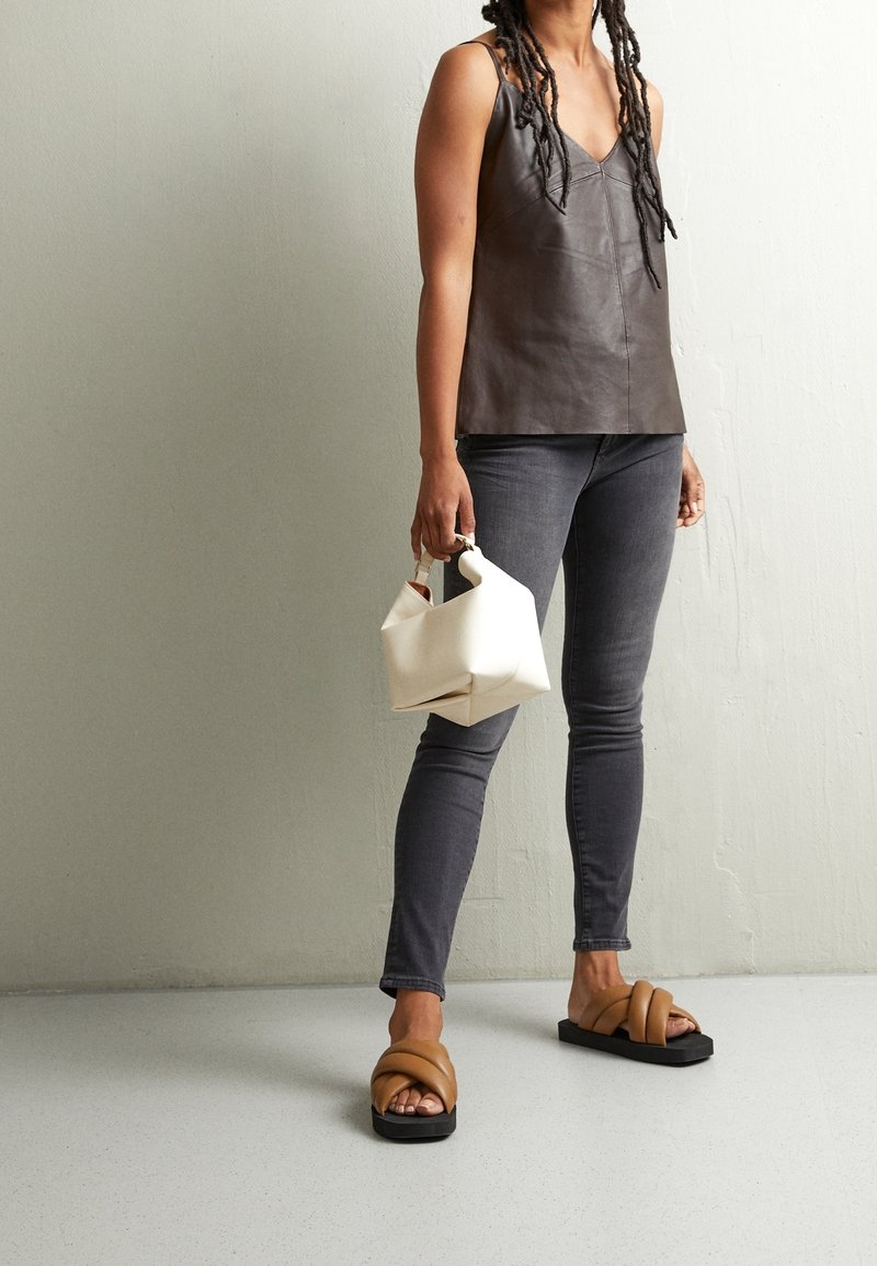 Top de cuero marrón con escote en V, jeans gris ajustados, sandalias negras con tiras cruzadas marrones, sosteniendo un bolso estructurado blanco.