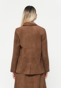 Veste en daim marron avec un col cranté et des manches longues, présentant une texture lisse et une fente simple à l'arrière.
