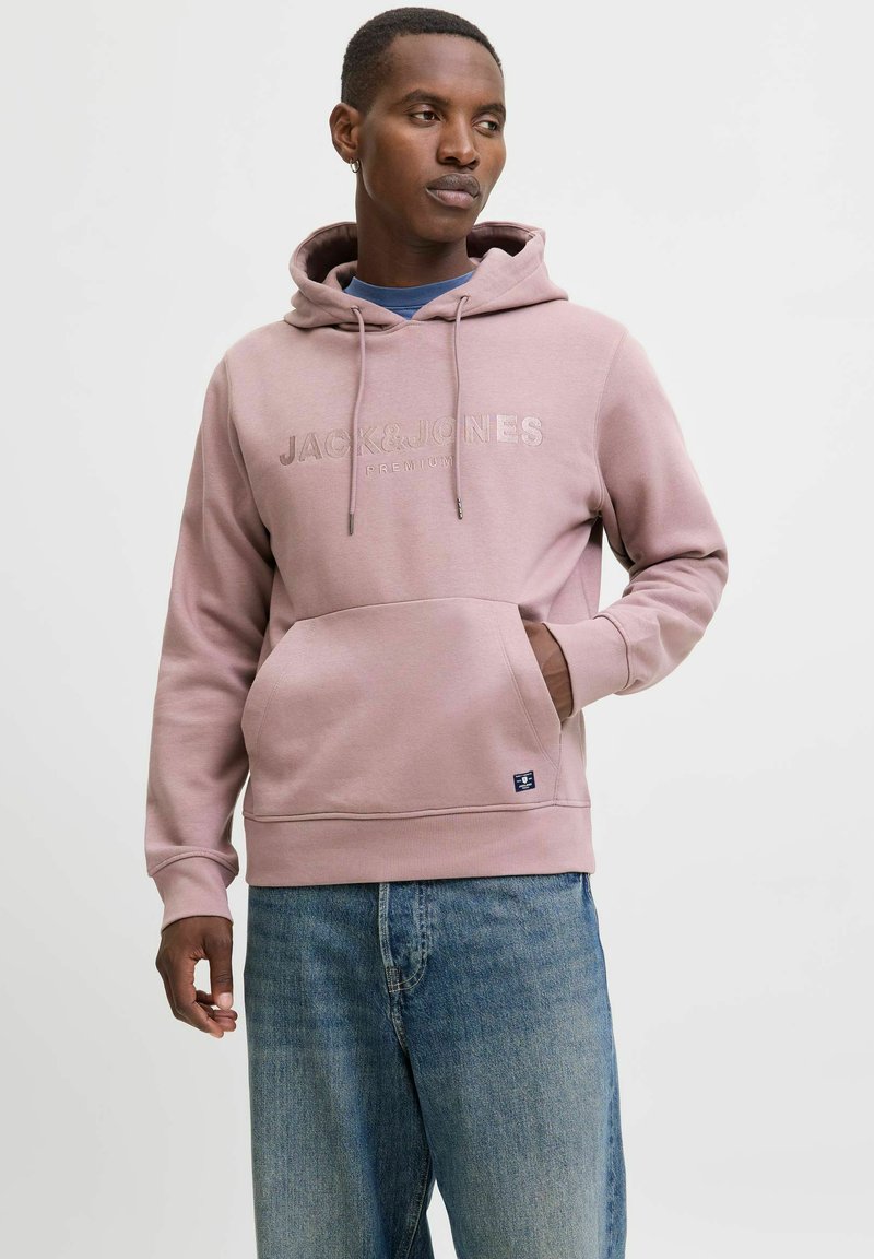 Hellrosa Hoodie mit Kängurutasche, Kapuze mit Zugband und geprägtem Logo. Mit gerippten Bündchen und Saum, kombiniert mit blauen Jeans.