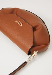 Wristlet clutch od teksturirane smeđe kože s zlatnim logotipom "GUESS EST 1981" i odvojivom trakicom na svijetloj pozadini.