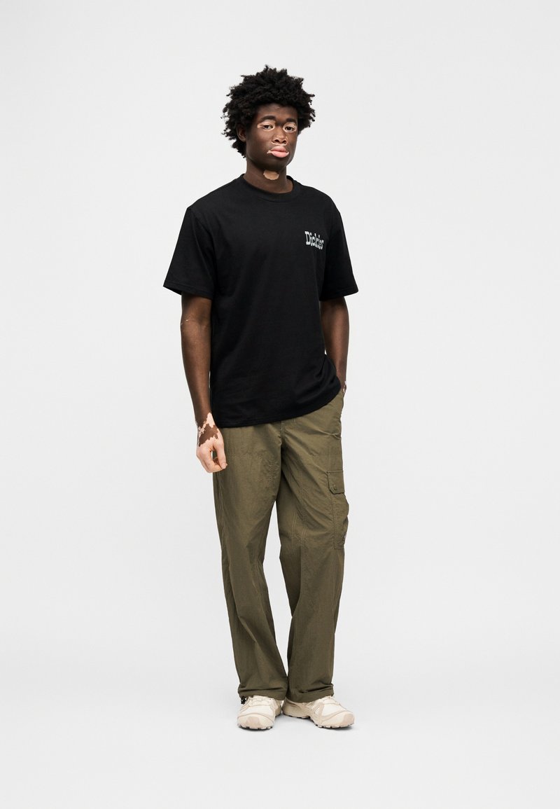 Black cotton t-shirt with logo, paired with olive green cargo pants. Model stands wearing cream-colored sneakers. Simple, casual outfit.