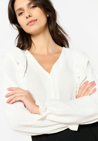 White blouse with a textured fabric, featuring a deep V-neck, ruffled shoulders, and button closures, paired with black attire.