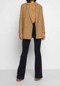 Tan double-breasted blazer over a beige button-up shirt, paired with dark flared jeans and black strappy sandals. Smooth fabric texture.