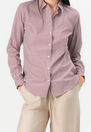 Kobieta ubrana w czerwoną i białą pasiastą koszulę na guziki z długim rękawem oraz beżowe spodnie, z rękami swobodnie w kieszeniach, na jednolitym tle.