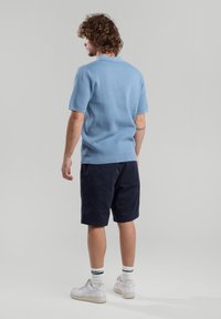 Light blue short-sleeve knit polo with a textured finish, paired with navy shorts and white sneakers, standing against a gray backdrop.