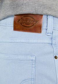 Jean en denim bleu clair avec une étiquette en cuir marron de la marque Dickies. Comprend des coutures le long des poches et un détail de bouton en laiton.