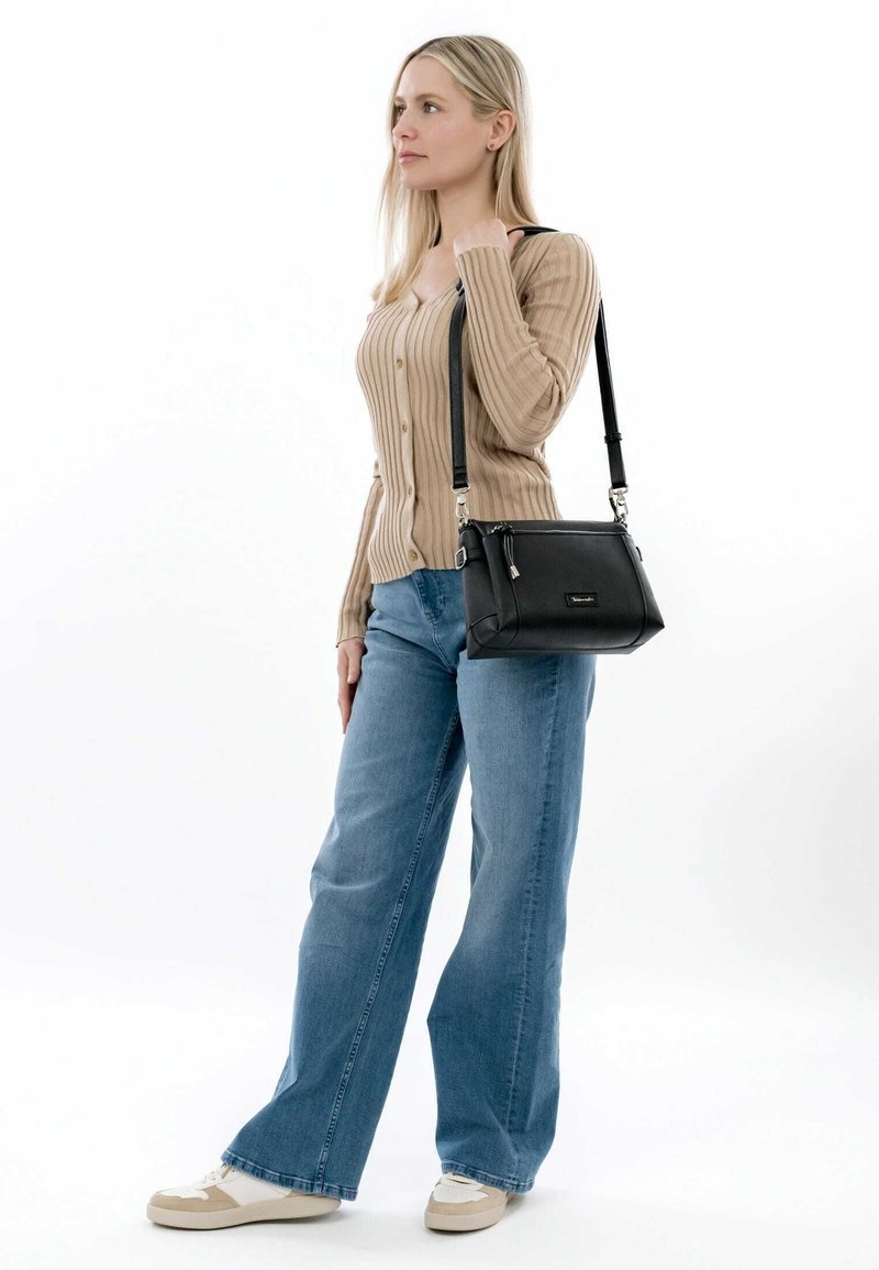 Black leather shoulder bag with silver hardware, rectangular shape, and adjustable strap. Model wears a ribbed beige sweater and blue jeans.