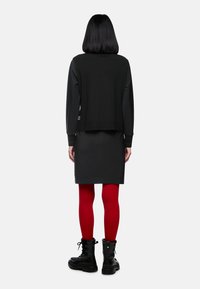 Person mit glattem schwarzen Haar steht mit dem Rücken zugewandt, trägt eine schwarze Jacke, einen schwarzen Rock, knallrote Strumpfhosen und schwarze Kampfstiefel.
