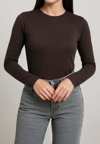 Top a maniche lunghe marrone scuro, dalla vestibilità aderente, con scollo rotondo e texture liscia, abbinato a jeans grigi a vita alta.
