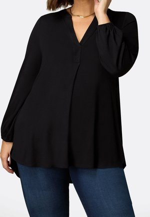 Vrouw met een losse zwarte lange mouw V-hals top met gerimpelde manchetten en blauwe jeans, poseert met één hand dichtbij het gezicht.