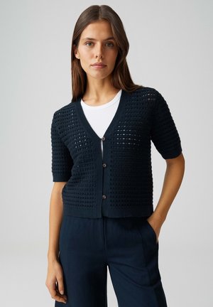 Vrouw draagt een marineblauwe gebreide korte-mouw cardigan over een wit shirt en marineblauwe broek, staand met één hand in de zak.